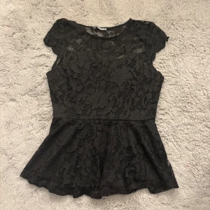 Svart spetsblus från Gina Tricot - Supersnygg svart blus i spets från Gina Tricot med blommigt mönster. Modellen har korta ärmar, rund hals och en peplum-nederdel som ger en feminin siluett. Perfekt att styla med jeans eller kjol för en trendig look.