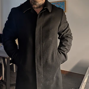 Svart klassisk burberryrock med rutig krage - Säljer en svart rock med stilren design och dold knäppning. Kragen har ett snyggt rutigt mönster på insidan som syns när du viker upp den. Jackan har långa ärmar och raka linjer, perfekt för dig som gillar en clean och tidlös look. Nypris; 20000 kr