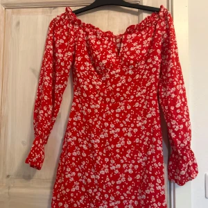 Röd blommig offshoulder-klänning Parisian - Superfin röd kortklänning från Parisian med vita blommor, offshoulder och långa ärmar. Klänningen har en lätt utställd form och smockad kant upptill. Perfekt för dig som vill sticka ut med en färgglad och söt look! Aldrig använd ettikett finns kvar 