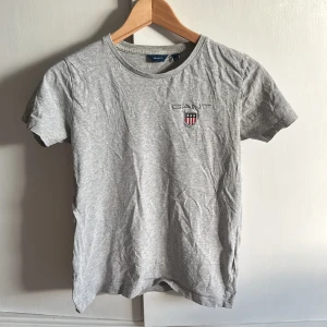 Grå t-shirt från GANT med logga - Snygg grå t-shirt från GANT med broderad logga och sköld på bröstet. Klassisk rund halsringning och korta ärmar. Tillverkad i mjuk bomull som känns skön mot huden. Perfekt för en clean och stilren look.  Använd men Inget att anmärka på T-shirten. 