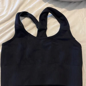 Svart ribbad sporttopp från Tridri - Svart ribbad sporttopp från Tridri med breda axelband och racerback. Toppen är croppad och har en tight passform som sitter skönt vid träning. Tillverkad i stretchigt syntetmaterial som andas och ger bra stöd.