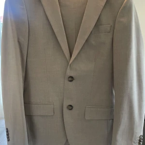 Beige kostym med kavaj och byxa - Stilren beige kostym med klassisk kavaj och matchande byxa. Kavajen har två knappar, smala slag och fickor med lock. Passformen är slim och tyget är lätt, perfekt för ljusare säsonger. Snygg och enkel design som ger en fräsch look.