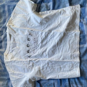 Vit t-shirt med månfas-tryck Hollister - Vit t-shirt från Hollister med tryck av månens faser på bröstet och diskret Hollister-logga. Klassisk rund hals och korta ärmar. Perfekt för dig som gillar clean stil med en twist. Mjuk och skön bomullskvalitet.