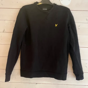 Svart sweatshirt från Lyle & Scott - Svart sweatshirt från Lyle & Scott med klassisk rund hals och gul broderad logga på bröstet. Tröjan har långa ärmar och ribbade muddar vid ärmslut och nederkant. Perfekt för en stilren och enkel look.