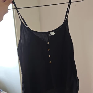 Svart linnetopp från H&M Divided - Svart linnetopp från H&M Divided med tunna, justerbara axelband och fyra små knappar framtill. Toppen är ärmlös och har en enkel, stilren look som passar perfekt till jeans eller kjol. Lätt och luftig känsla, perfekt för varma dagar.