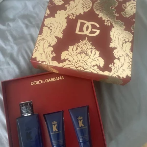 Dolce & Gabbana K parfymset - Lyxigt parfymset från Dolce & Gabbana med doften K. Innehåller en blå glasflaska med silverkrona som lock samt två matchande blå tuber. Allt levereras i en elegant röd och guld ask med dekorativt mönster och DG-logga.