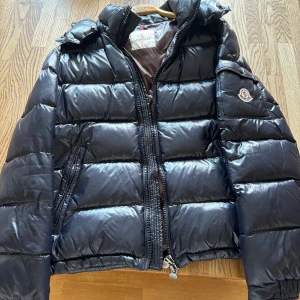 Moncler Maya jacka - Snygg  maya från Moncler med glansig finish och klassisk logga på ärmen. Jackan har huva, dragkedja framtill och praktiska fickor. Perfekt för kalla dagar när du vill hålla stilen och värmen.