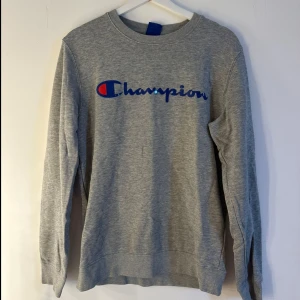 Grå Champion sweatshirt i bomull - Snygg grå sweatshirt från Champion med klassisk logga i blått och rött framtill. Tröjan har rund halsringning och långa ärmar. Perfekt för en chill och sportig stil. Tillverkad i mjuk bomull för extra komfort. Storlek s