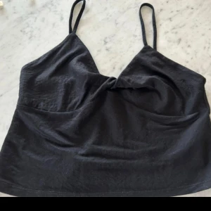 Svart linnetopp med twist framtill - Säljer en svart linnetopp med smala axelband och snygg twist-detalj framtill. Toppen har en enkel, croppad passform och är perfekt för att styla med jeans eller kjol. Mjuk och lätt i materialet, perfekt för varma dagar.