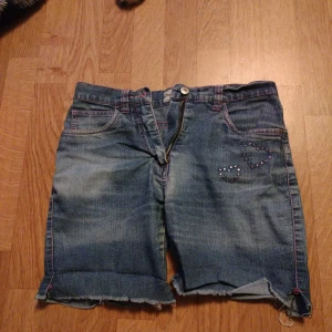 Jeansshorts med hjärtan och nitar - Snygga blå jeansshorts med råa kanter och uppvik. Dekorerade med hjärtan av nitar både fram och bak. Klassisk femficksmodell med dragkedja och knapp. Perfekta för sommaren och ger en cool, avslappnad vibe.
