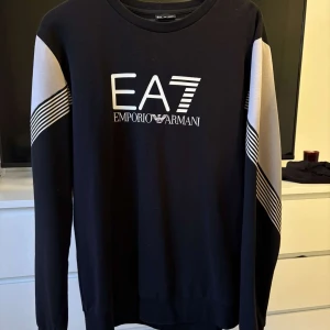Svart EA7 Emporio Armani sweatshirt XL - Svart långärmad sweatshirt från EA7 Emporio Armani med vitt tryck framtill och vita paneler med svarta ränder på ärmarna. Klassisk rund halsringning och relaxed fit. Perfekt för dig som gillar stilren streetwear med premiumkänsla.