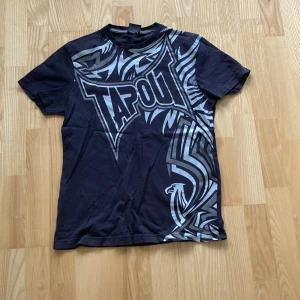 Svart Tapout t-shirt med tryck - Cool svart t-shirt från Tapout med stort grafiskt tryck i silvergrått över framsidan och en örn-detalj. Klassisk passform med rund hals och korta ärmar. Perfekt för dig som gillar streetwear och sportiga vibes.