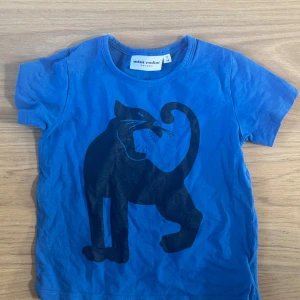 Blå t-shirt med pantermotiv Mini Rodini - Blå t-shirt från Mini Rodini med ett stort svart pantermotiv på framsidan. T-shirten har rund halsringning och korta ärmar. Tillverkad i mjuk ekologisk bomull som känns skön mot huden. Perfekt för dig som gillar coola prints och färg.