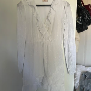 Vit omlottklänning med volang från Nelly - Superfin vit kort omlottklänning från Nelly med volangdetaljer längs v-ringningen och nederkanten. Klänningen har långa ärmar, knytband i midjan och ett diskret prickigt mönster i tyget. Använd en gång.