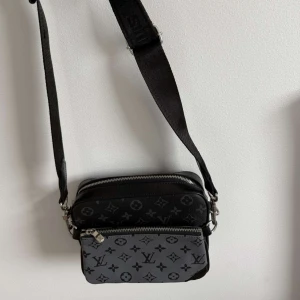  axelväska monogram - Snygg svart axelväska  med klassiskt monogrammönster i grått och svart. Väskan har två fack med dragkedja och justerbar. Perfekt för att bära det viktigaste och ge din outfit en lyxig touch.