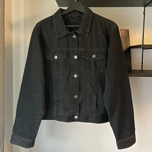 Svart jeansjacka  - Snygg svart jeansjacka med klassisk krage och silverfärgade knappar framtill. Jackan har två bröstfickor med lock och knapp, samt långa ärmar med knapp vid ärmslut. Passar perfekt till dig som är mellan ca 170-180cm. 