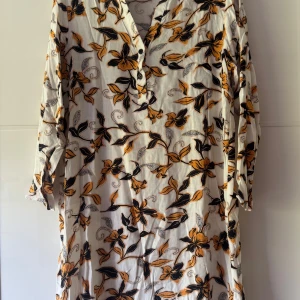 Blommig tunika/klänning från Dea - Tunika/kläning från Dea i vitt med blommigt mönster i gult och svart. Klänningen har långa ärmar, v-ringning med knappar och är tillverkad i ett lätt och luftigt material, perfekt för varma dagar. Snygg och avslappnad passform med bohemisk känsla.