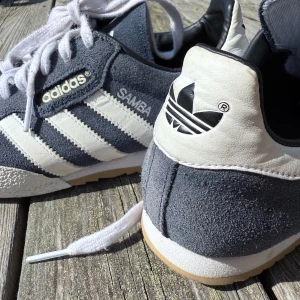 Retro Adidas Samba sneakers i blå och vitt - Ett par unika Adidas Samba sneakers i mörkblå mocka med vita läderdetaljer och de ikoniska tre ränderna på sidorna. Skorna har vit snörning, gummisula och svart Adidas-logga på hälen. Perfekta för dig som gillar retrostil och streetwear. 