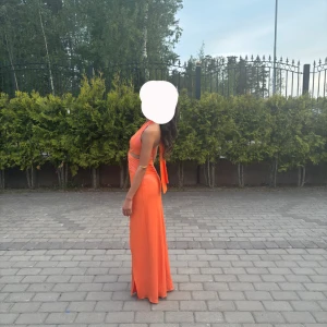 Orange lång klänning med slits - Snygg orange långklänning med halterneck och öppen rygg. Klänningen har en twistad detalj framtill, markerad midja, hög slits framtill och glimmrar i tyget. Mjuk och följsam i materialet, perfekt för bröllop eller bal. Pris går även att diskutera