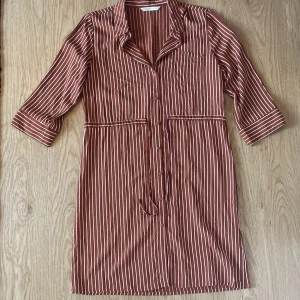 Randig skjortklänning från ONLY storlek 34 - Snygg skjortklänning från ONLY i brun med vita vertikala ränder. Modellen har trekvartsärm, knappar hela vägen framtill, krage och två bröstfickor. Midjeband för att knyta och markerad midja. Perfekt för en chill och trendig look.                                     OBS! Stygn har släppt på ett par cm på midjebandet som syns på bild 4.