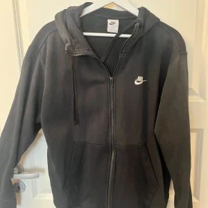 Svart Nike hoodie med dragkedja - Svart hoodie från Nike med vit logga på bröstet. Tröjan har huva med snörning, hel dragkedja och två fickor framtill. Perfekt för chill dagar eller när du vill ha en sportig look. Tillverkad i mjukt material för extra komfort.