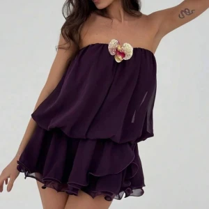 Mörklila axelbandslös volangklänning - Superfin mörklila kortklänning med axelbandslös design och volanger i flera lager. Klänningen har en unik blomdetalj framtill och luftig, flowy passform.