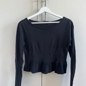Svart långärmad topp med volang - En svart långärmad topp med rund halsringning och volangdetalj nedtill. Modellen är croppad och har en enkel, stilren look som passar perfekt till jeans eller kjol. Tillverkad i mjukt bomullstyg för en bekväm känsla hela dagen.