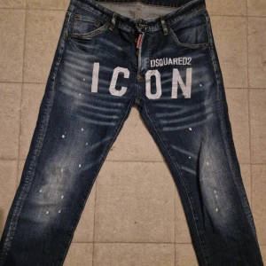 Dsquared2 ICON jeans med tryck - Snygga blå jeans från Dsquared2 med stort vitt 'ICON' tryck och DSQUARED2-logga framtill. Jeansen har en tvättad look med slitningar och vita färgstänk för en cool streetstyle. Klassisk femficksmodell och normal passform. Perfekta för dig som vill sticka ut. Pris kan diskuteras