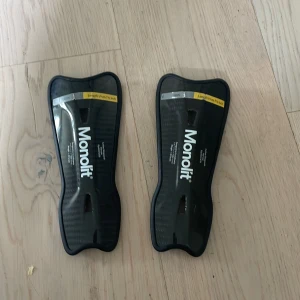 Monolit benskydd med förpackning  - Säljer ett par svarta Monolit benskydd 19 cm med förpackning . De är lätta och har en snygg, modern design med ventilerande hål och en skyddande yta i plast. Kommer med ett praktiskt fodral för enkel förvaring och transport.