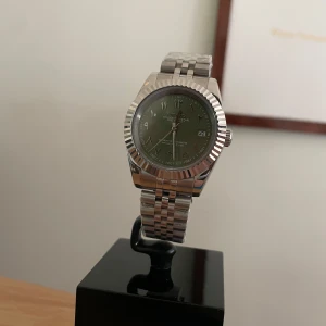 Seiko Mod Olive green urtavla med arabiska siffror - Snygg Seiko automatisk klocka med grön urtavla och datumvisning. Boetten och länken är i blankt silverfärgat stål med räfflad bezel och klassisk länkdesign. Urtavlan har arabiska siffror och tydliga visare. Perfekt accessoar för dig som gillar stilrena detaljer. Äkta NH35 Urverk! 5ATM Rostfritt Stål