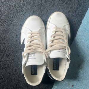 Vita sneakers från Superga med svart detalj - Snygga vita sneakers från Superga med svart stjärndetalj på sidan. Skorna har klassisk låg profil, beige snörning och platt sula. Ovandelen är i skinn och detaljer i mocka vid hälen. Perfekta för en clean och stilren look.