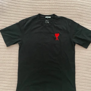 Svart t-shirt från AMI med rött hjärta - Svart t-shirt från AMI Alexandre Mattiussi med klassisk passform. På bröstet finns ett broderat rött hjärta med bokstaven A. T-shirten har rund halsringning och är tillverkad i mjuk bomull. Perfekt för dig som gillar stilrena plagg med en twist.