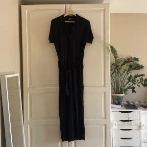 Svart långklänning Dr. Denim - Svart långklänning från Dr. Denim med korta ärmar, krage och knappar framtill. Klänningen har ett knytband i midjan och är tillverkad i mjuk bomull. Enkel och stilren design som passar perfekt till många tillfällen. Storlek XS, passar upp till M. I fint skick.