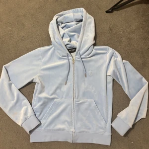 Ljusblå velour hoodie Juicy Couture - Supermjuk ljusblå hoodie från Juicy Couture i velour med dragkedja framtill, huva och fickor. Klassisk JC-logga broderad på bröstet. Perfekt för en chill och trendig look.