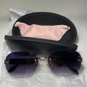 Svarta Miu Miu solglasögon med lila glas - Snygga solglasögon från Miu Miu med svarta, breda skalmar och guldiga detaljer. Glasen är lila-tonade och har en cool, kantig form. Kommer med originalfodral och putsduk. Perfekta för dig som vill sticka ut med en lyxig vibe.