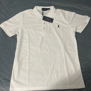 Vit pikétröja Polo Ralph Lauren - Klassisk vit pikétröja från Polo Ralph Lauren med broderad logga på bröstet. Tröjan har korta ärmar, krage och knäppning med tre knappar framtill. Slim fit-modell i bomull som ger en fräsch och stilren look. (OBS! Tröjan är inte autentisk)