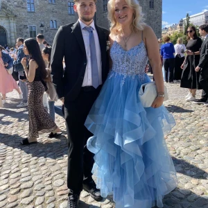 Blå långklänning med spets och volanger - Drömmig blå långklänning med lager på lager av volanger i kjolen och spetsdetaljer upptill. Klänningen har smala axelband, v-ringning och snörning i ryggen. Perfekt för bal eller student. Materialet är lätt och ger en riktigt prinsessig känsla. Köpte för 3300kr