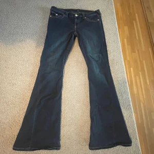 True Religion Joey Low Rise Flare Jeans - Säljer ett par mörkblå True Religion Joey jeans med låg midja och rejäl bootcut flare. Klassiska kontrastsömmar, snygga detaljerade bakfickor med knappar och logga. Tillverkade i jeansmaterial, perfekta för dig som gillar statement-jeans med 2000-talsvibe.