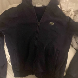Mörkblå fleecejacka från Lacoste - Mörkblå fleecejacka från Lacoste med klassisk krokodillogga på bröstet. Jackan har dragkedja framtill, ståkrage och två fickor. Perfekt för lager-på-lager och chill vardagsstil.