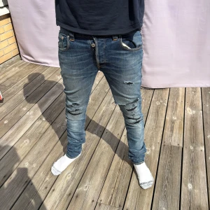 Blå slitna Nudie jeans  - Säljer ett par blå slim jeans med slitningar och detaljer på knäna. Nudie Jeansen har klassisk femficksdesign, orange sömmar och snygg tvätt. Passformen är tight och de sitter snyggt längs benen. Perfekta för dig som gillar en lite ruffare streetstil. Extremt bra skick men lite för tajta för mig! Pris kan diskuteras! 