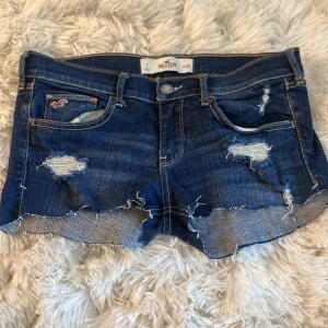 Blå slitna jeansshorts Hollister - Snygga blå jeansshorts från Hollister med råa kanter och slitningar framtill och på bakfickorna. Klassisk femficksmodell med låg midja och coola detaljer som ger en avslappnad vibe. Perfekta för sommardagar och festivaler. (((( Dom är klippt själv))))