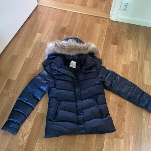 Mörkblå Moncler jacka med päls huvan äkta - Snygg mörkblå dunjacka från Moncler med fluffig fuskpäls på huvan. Jackan har inga skador passar väldigt bra på vintern har aldrig varit kall på vintern med den nu är det sommar men man behöver den på vintern köp snälla den behöver få sålt den 😀