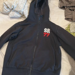 Svart hoodie Sail Racing SR77 - Svart hoodie från Sail Racing med dragkedja och huva. Broderad SR 77-logga i vitt och rött på bröstet. Mjuk insida och klassisk passform, perfekt för chill dagar. Snygga detaljer med röd kant på insidan av huvan.