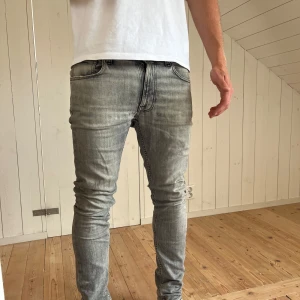 Grå jeans från Nudie Jeans - Säljer ett par grå skinny jeans från Nudie Jeans med slitningar och snygg tvätt. Jeansen är storlek 29, 32 och har  svarta detaljer och smal passform - lite för små för mig så kan sitta olika! Dom har tyvärr lite skavanker (kolla bild) - därmed bra pris/ eller bud! 