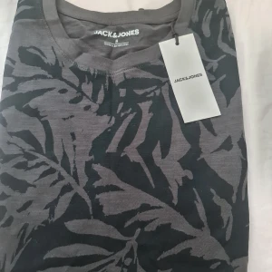 Mörkgrå mönstrad tröja Jack & Jones - Snygg mörkgrå tröja från Jack & Jones med svart bladliknande mönster. Tröjan har rund halsringning och är tillverkad i mjuk bomull, perfekt för dig som gillar en stilren men ändå unik look.
