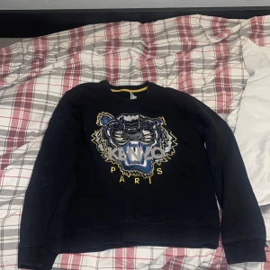 Svart Kenzo sweatshirt med tiger - Svart sweatshirt från Kenzo med ikoniskt broderat tigerhuvud i blått, grått och gult framtill. Tröjan har rund hals, långa ärmar och ribbade muddar. Perfekt statement-plagg för dig som gillar streetstyle och vill sticka ut.
