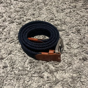 Mörkblått flätat bälte Jack & Jones - Snyggt mörkblått flätat bälte från Jack & Jones med detaljer i brunt skinn och silvrigt spänne. Bältet har en stilren look med klassisk design och passar perfekt till jeans eller chinos. Märket är präglat på skinnet vid spännet.