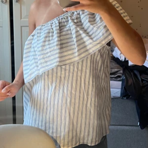 Randig oneshoulder topp från H&M - Säljer en luftig och somrig randig topp från H&M i oneshoulder-modell. Toppen är vit med tunna blå ränder och har en bred volang över axeln. Perfekt för varma dagar och enkel att styla med jeans eller kjol.