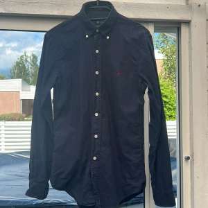 Mörkblå skjorta från Ralph Lauren - Snygg mörkblå skjorta från Ralph Lauren i slim fit-modell. Skjortan har klassisk krage, vita knappar och det ikoniska röda broderade Polo-loggan på bröstet. Tillverkad i mjuk bomull och med långa ärmar. Perfekt för en clean och stilren look.