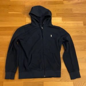 Svart hoodie från Ralph Lauren - Svart hoodie från Ralph Lauren med dragkedja och klassisk vit logga på bröstet. Tröjan har huva, två fickor framtill och långa ärmar med muddar. Perfekt för en chill och stilren look.